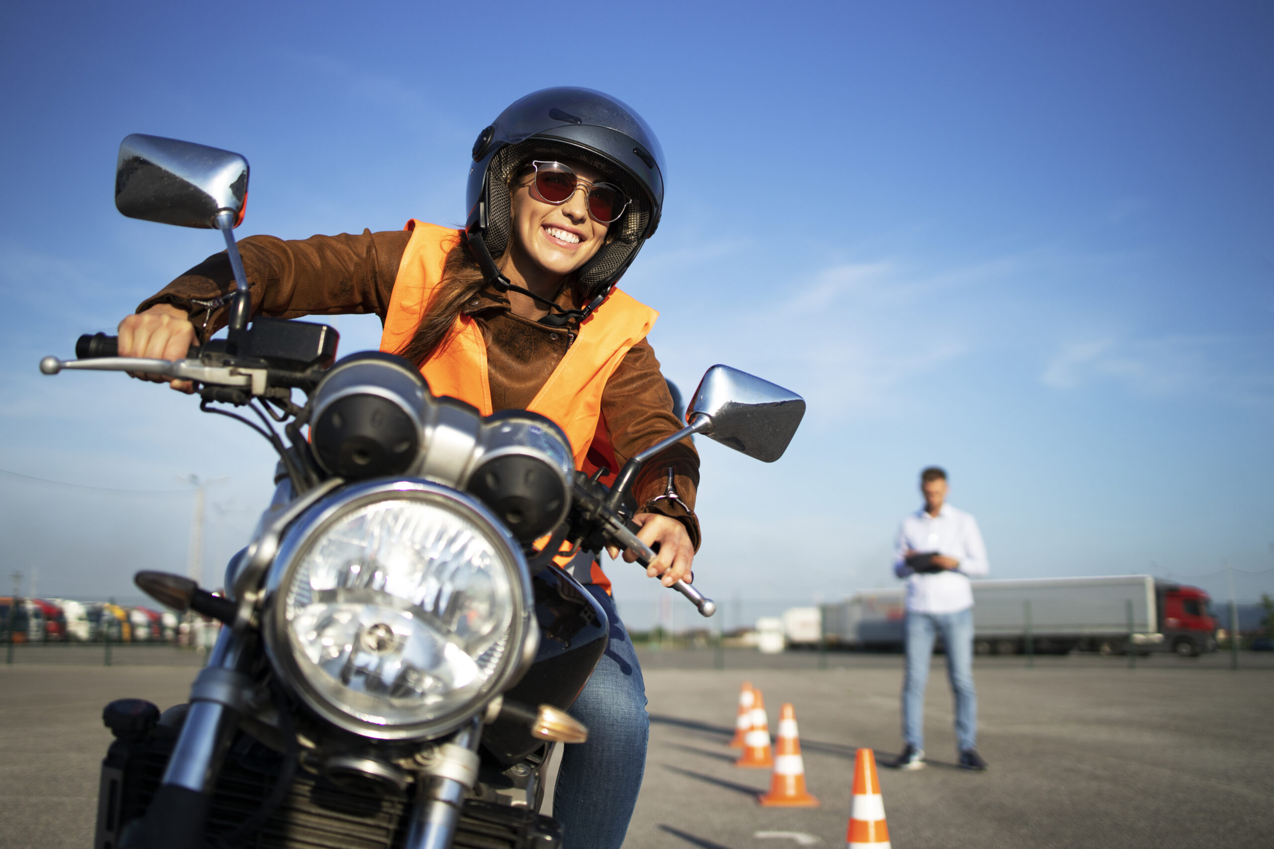 Female student with helmet taking motorcycle lessons and practicing ride.  In background traffic cones and instructor with checklist rating and  evaluating the ride. Motorcycle school of driving. - Mechanical Booster
